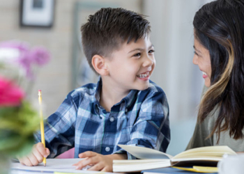 Adorable elementary age boy works on homework assignment. His mom is helping him with the assignment.