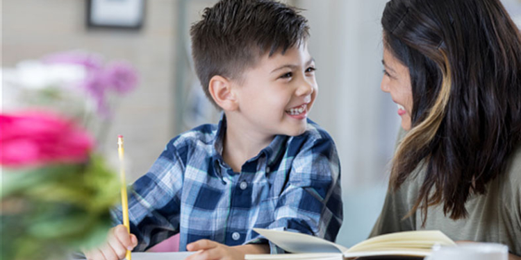 Adorable elementary age boy works on homework assignment. His mom is helping him with the assignment.